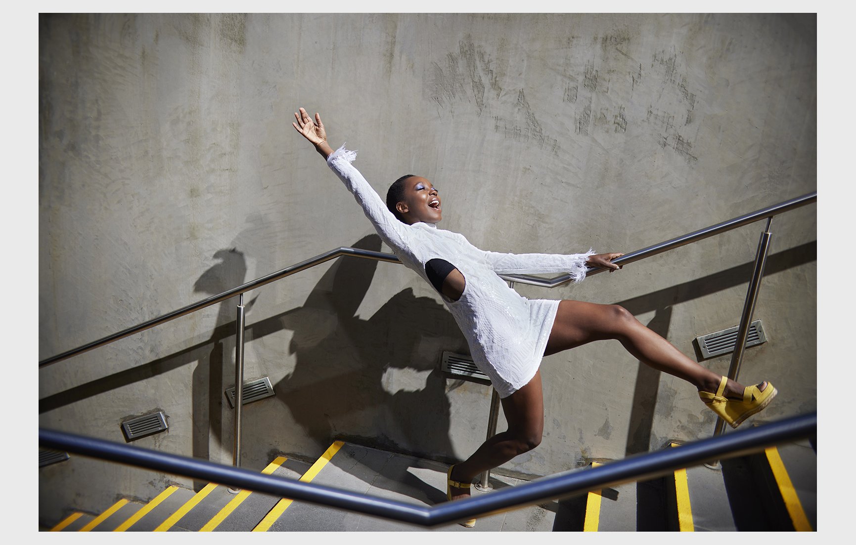 Woman poses in a white dress with arm raised on stairs with yellow stripes, in an indoor setting with a concrete wall backdrop.