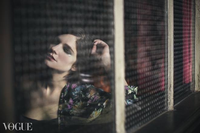 Indoor portrait of an adult woman in a floral jacket with hand in hair, looking out of an iron-grated window. Vogue logo visible.