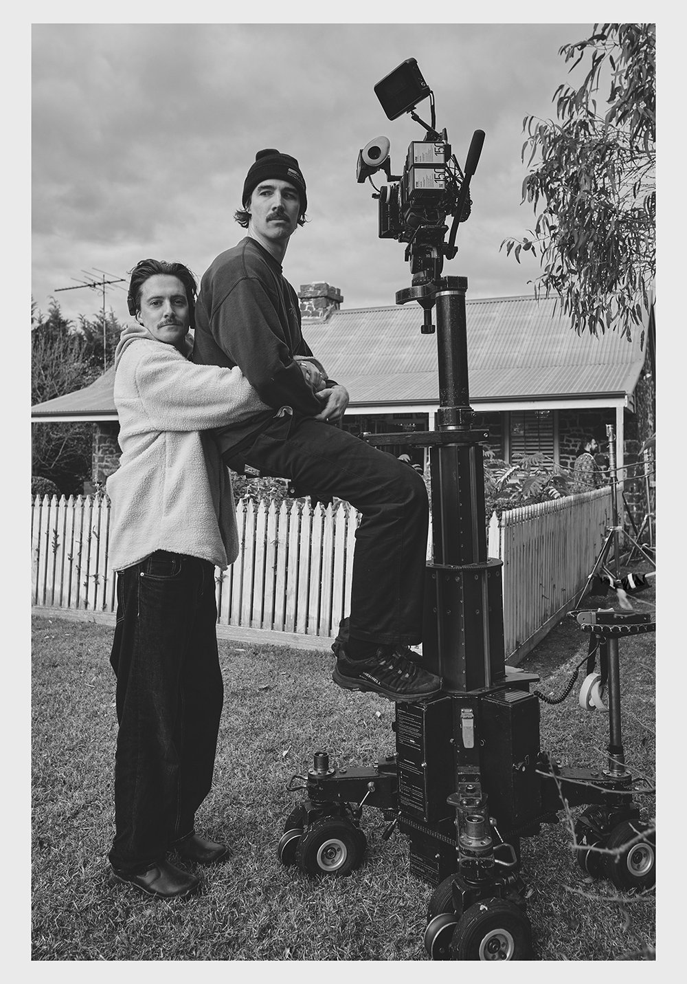 Monochrome shot of two adult men posing with a large film camera on a wheeled platform in a grassy yard, a house in the background.