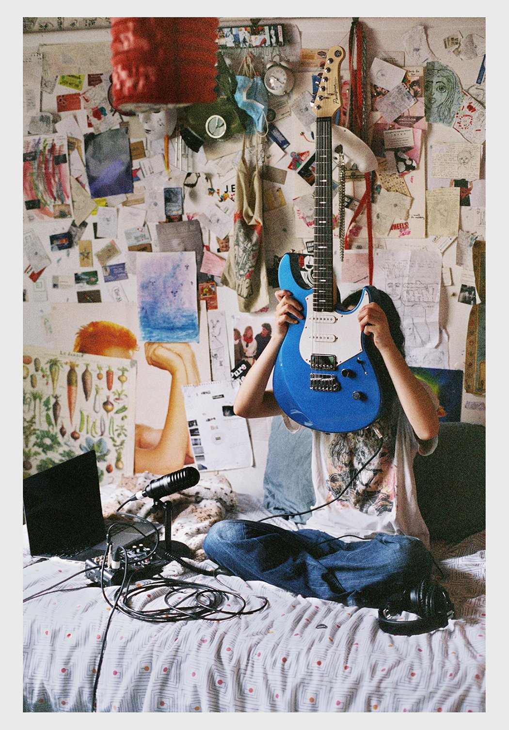 Teenager sits cross-legged on a bed, holding a blue electric guitar in front of face, in a room filled with papers.