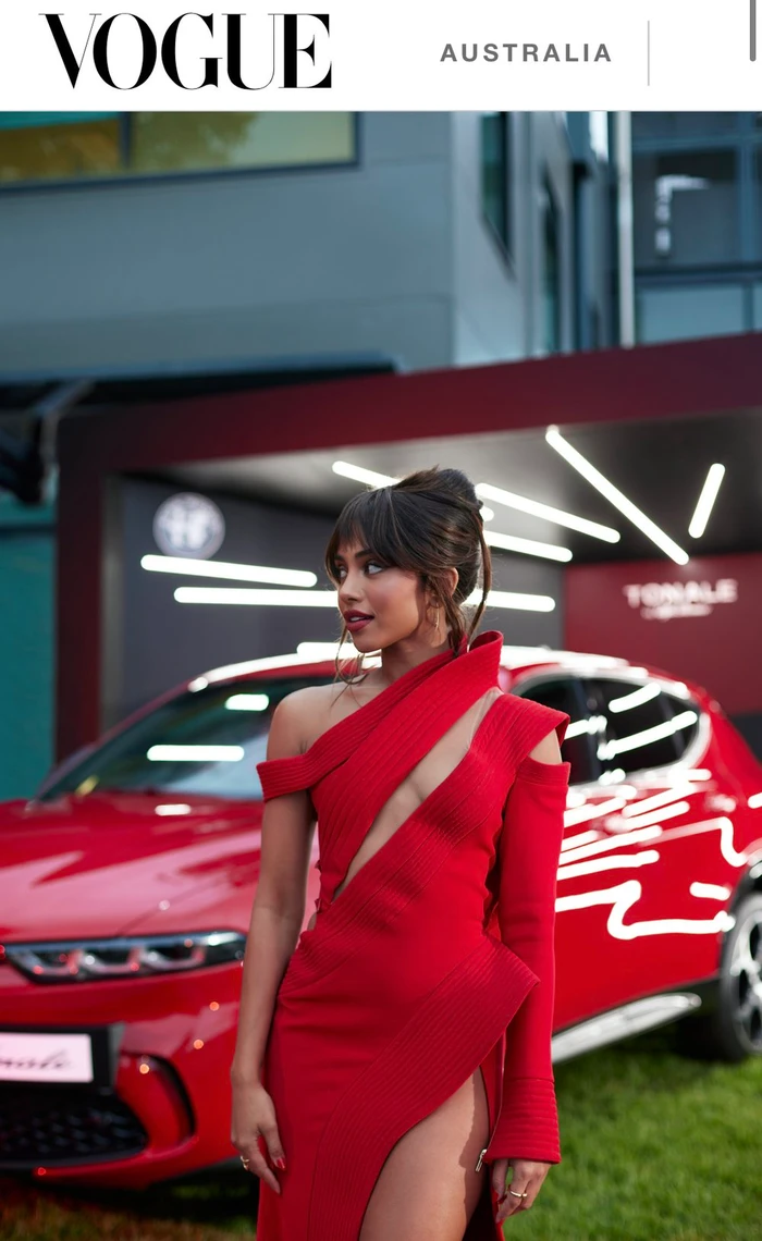 Woman wearing a red dress standing in front of a red car outdoors with Vogue Australia branding at the top.