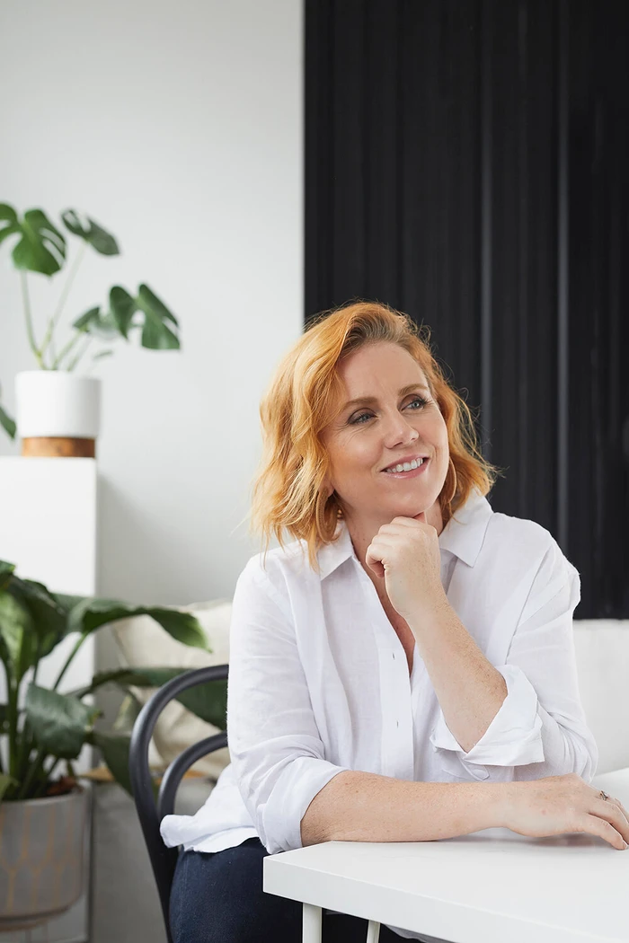 Woman with shoulder-length reddish-blonde hair wearing a white collared shirt seated at a white table in a bright interior