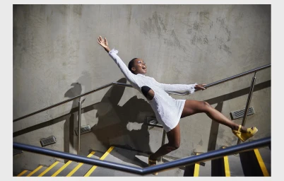 A Black woman in a white mini dress leaning backward against a metal railing on outdoor stairs in Melbourne by Catherine Black