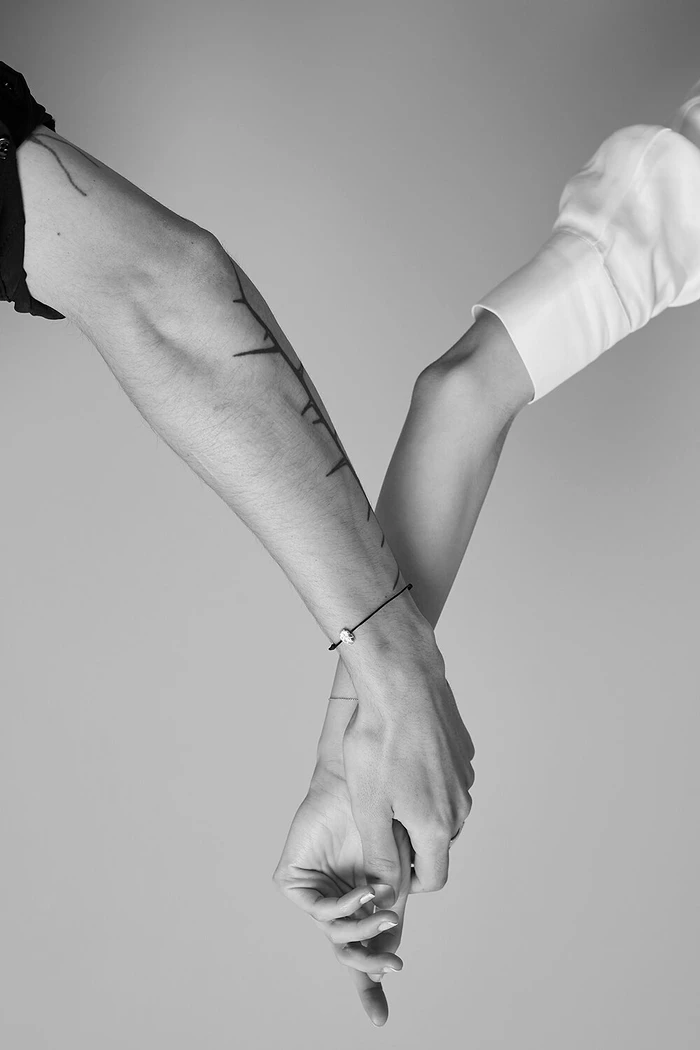 Monochrome close-up of two people holding hands against a neutral background, featuring tattooed arms and a delicate bracelet.
