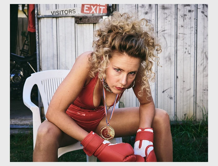 Blonde woman wearing a red vinyl swimsuit and boxing gloves with a gold medal seated outdoors on a white plastic chair