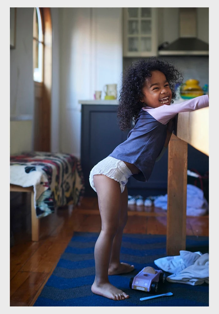 Young child with curly hair laughing and leaning against a wooden table in a domestic kitchen by Catherine Black