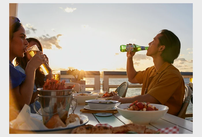 People dining outdoors at sunset on a rooftop overlooking water while drinking beer and eating seafood.