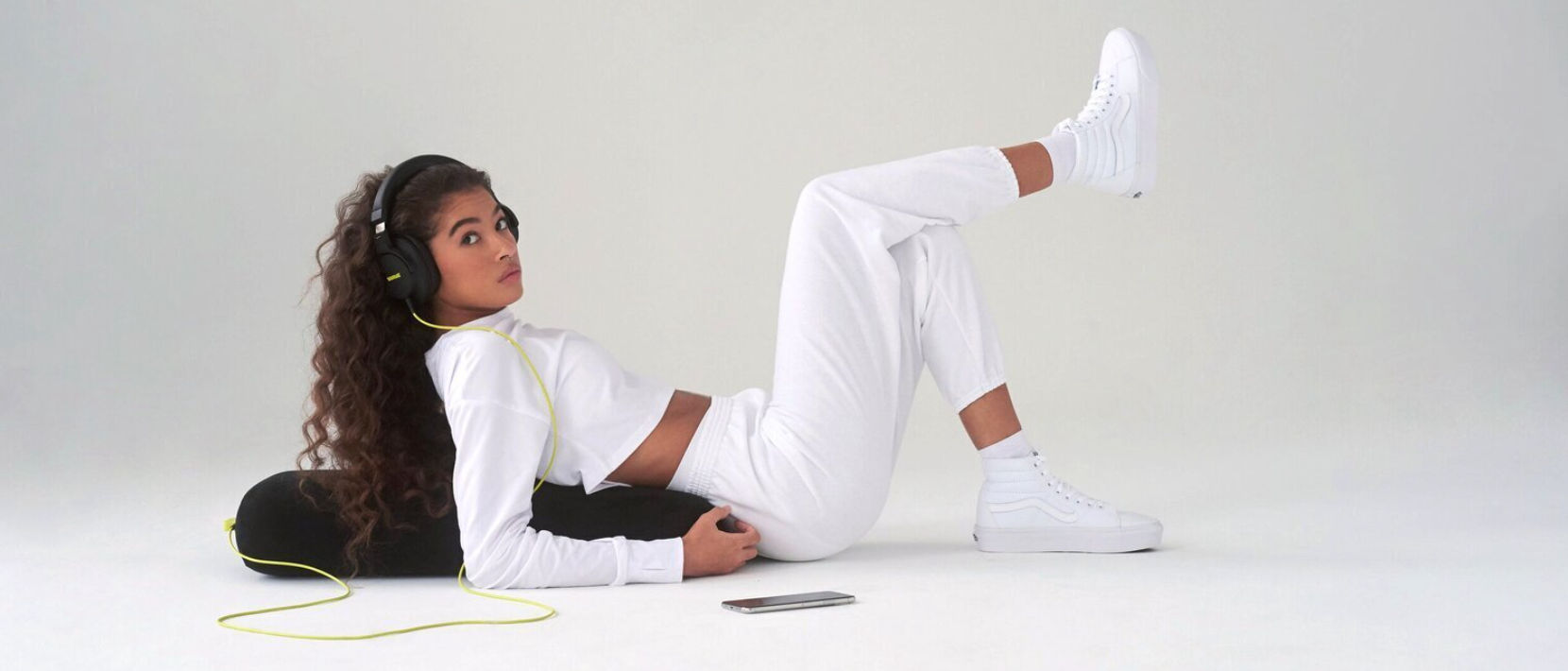 Young woman wearing headphones reclines in an all-white outfit against a grey background with a black cushion and mobile phone.