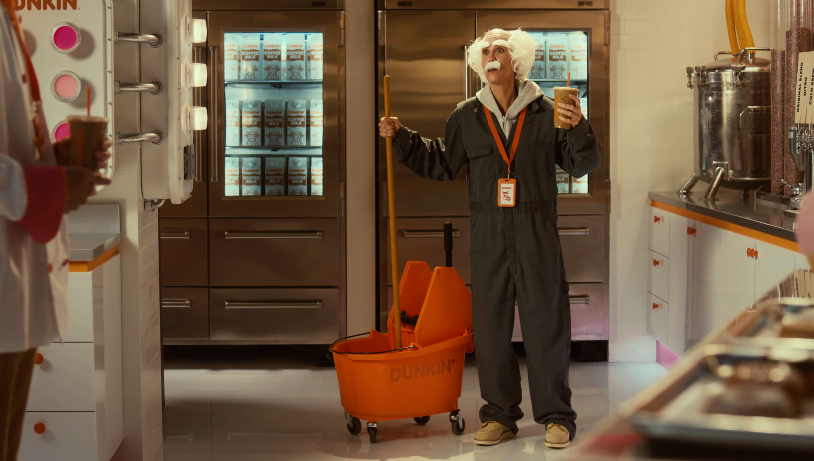 Man in a gray jumpsuit, white wig and mustache stands in a Dunkin' kitchen holding a coffee drink and a mop with a mop bucket.