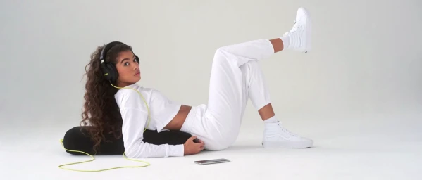 Young woman in white loungewear and white sneakers reclining against a black bolster pillow wearing black headphones.