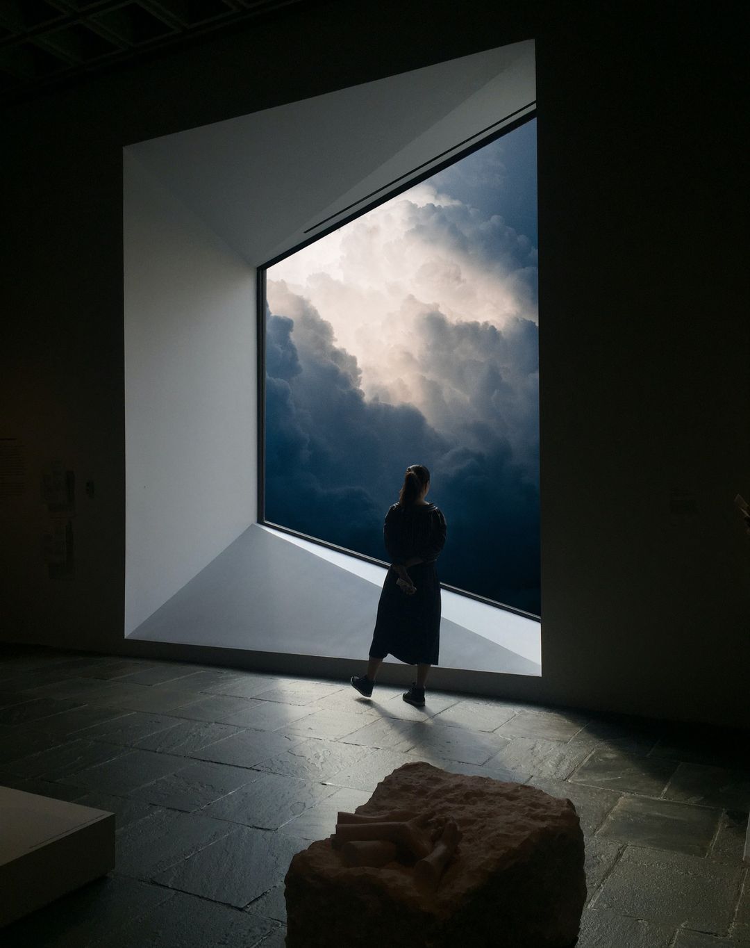 Person silhouetted in modern art gallery stands near window overlooking stormy sky; monolithic sculpture in foreground.