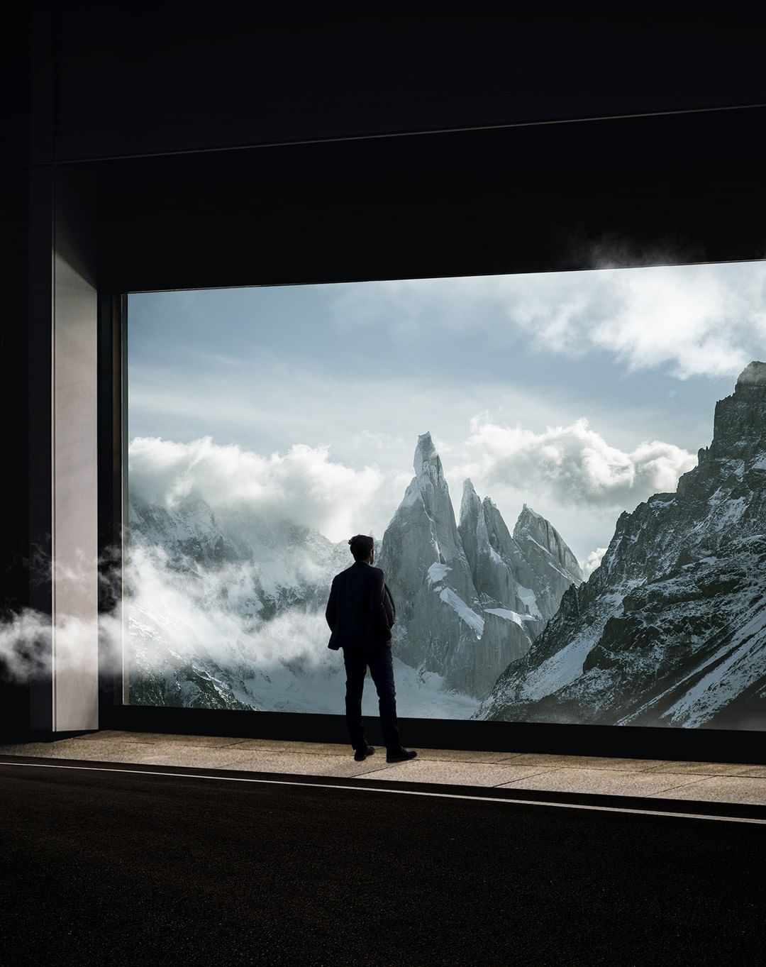 A solitary man stands before a window showcasing a majestic mountain range partially shrouded in clouds, set against a dimly lit foreground.