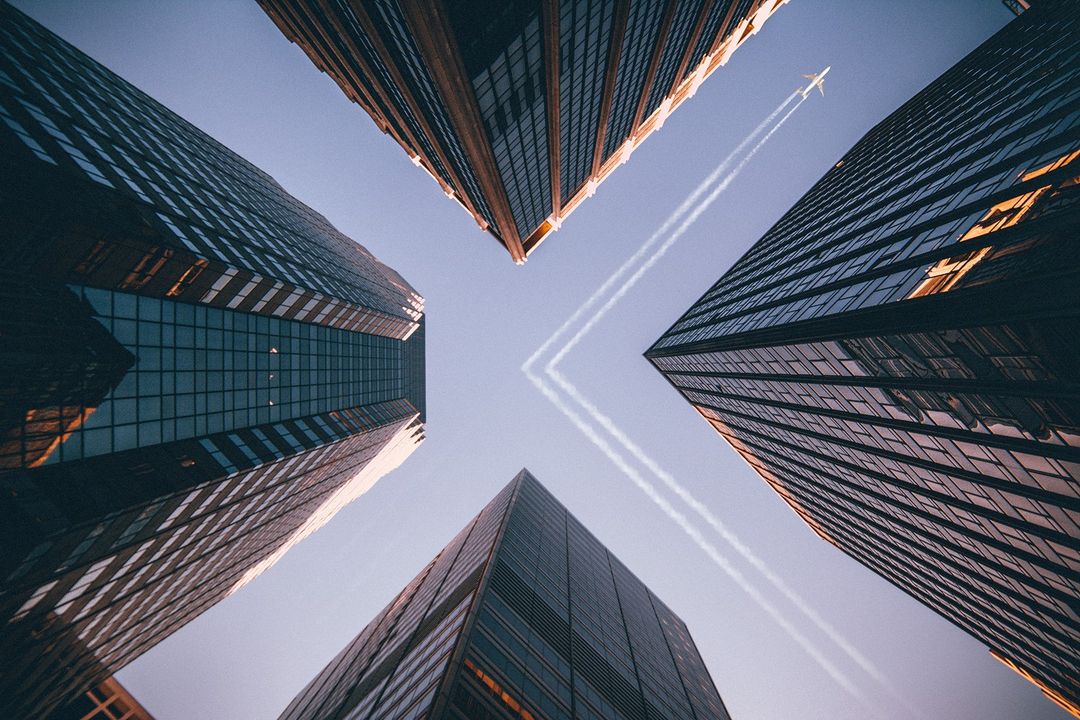 Low angle view of skyscrapers reaching toward the sky with airplane contrails above, creating a modern urban landscape.