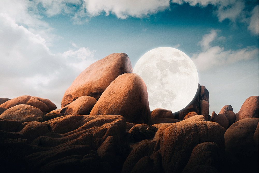 Full moon behind rock formations with a partly cloudy blue sky in the background, creating a landscape with a natural, celestial focus.