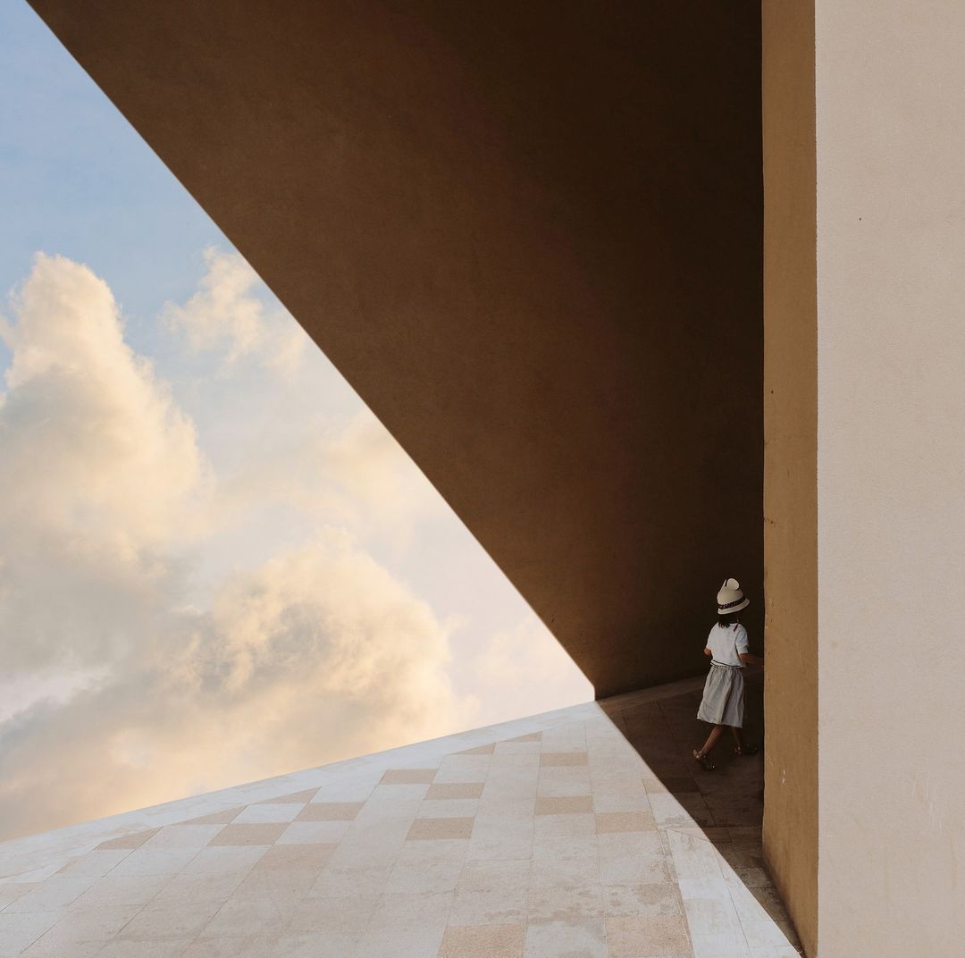 Young girl in sun hat walks near modern tan-colored architectural structure against a partly cloudy blue sky in an outdoor setting.