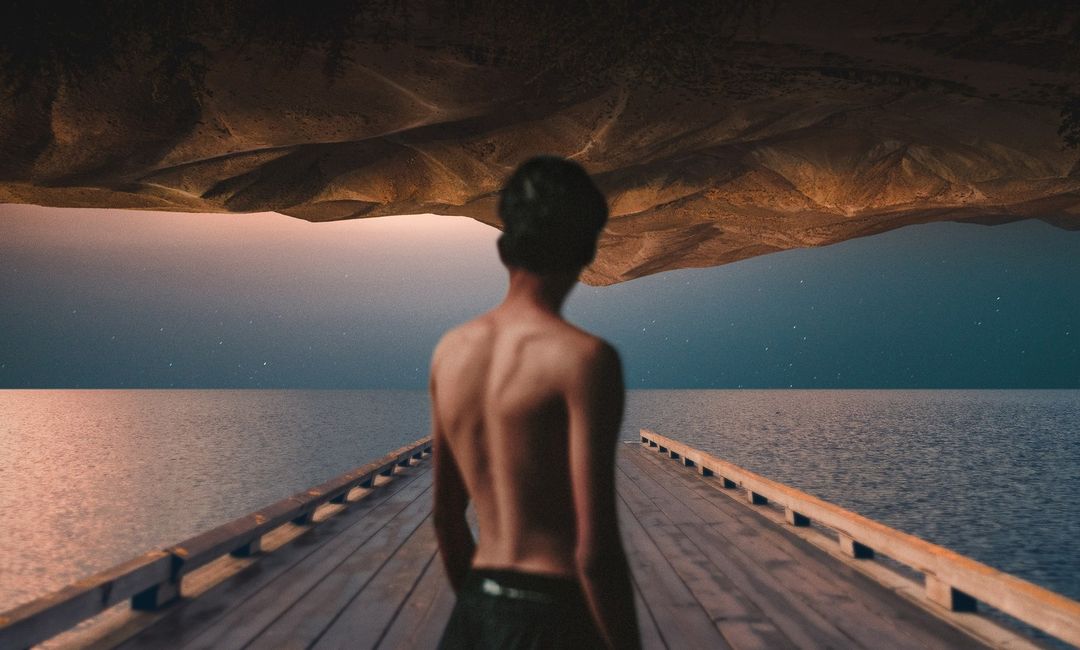 Man stands on a wooden dock, facing a tranquil ocean under a dusky sky with an upside-down mountain looming overhead in a surreal scene.