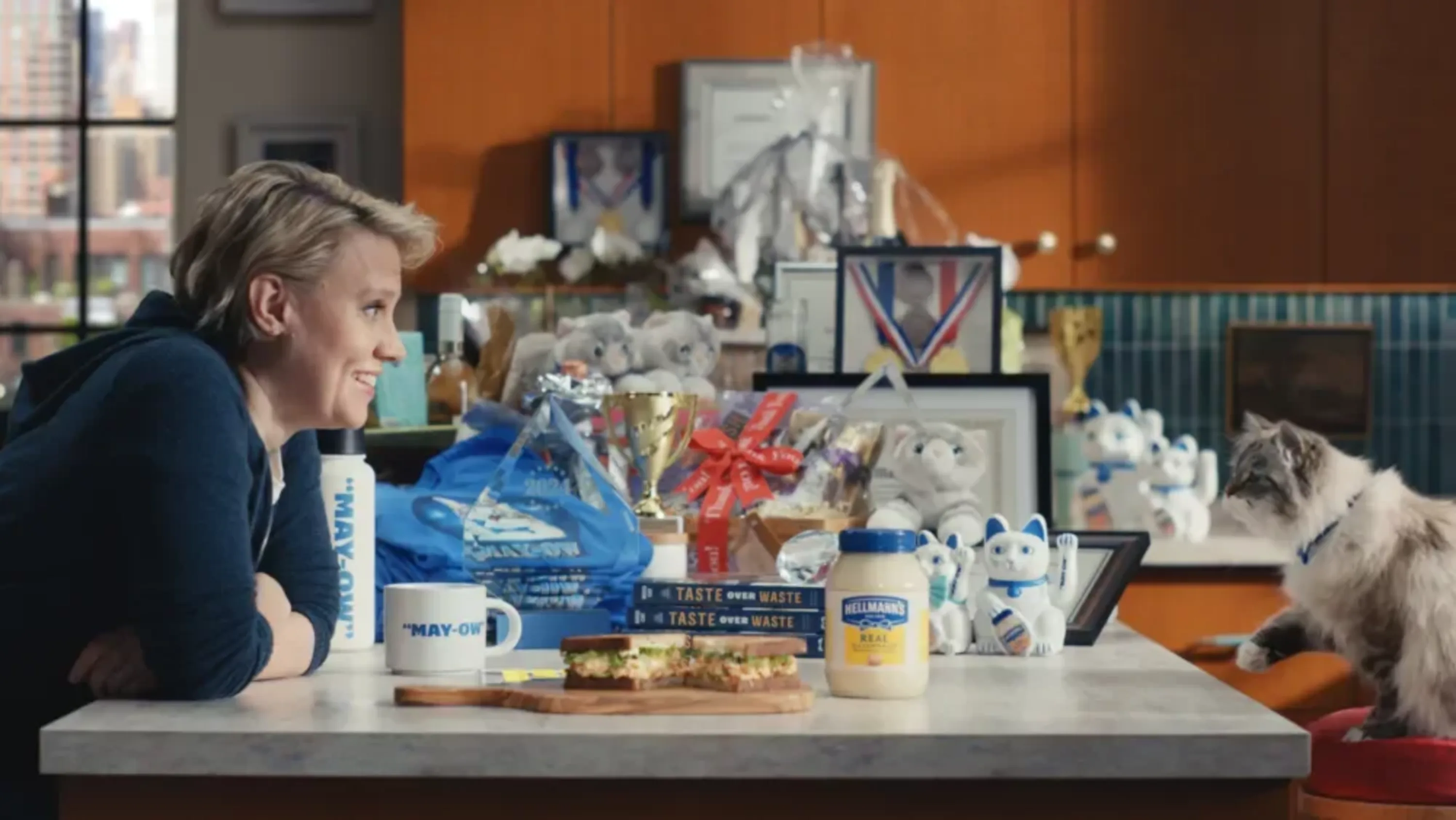 Woman at a kitchen counter with a cat, food, and Hellmann's mayonnaise, enjoying a sandwich in a cozy, bright indoor setting.