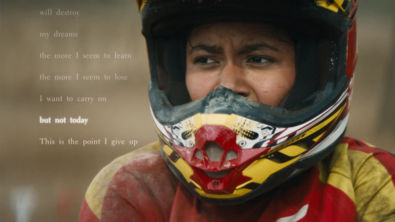 Close-up of a person wearing a motocross helmet and red jersey, looking to the side with a determined expression, set against a blurred background.