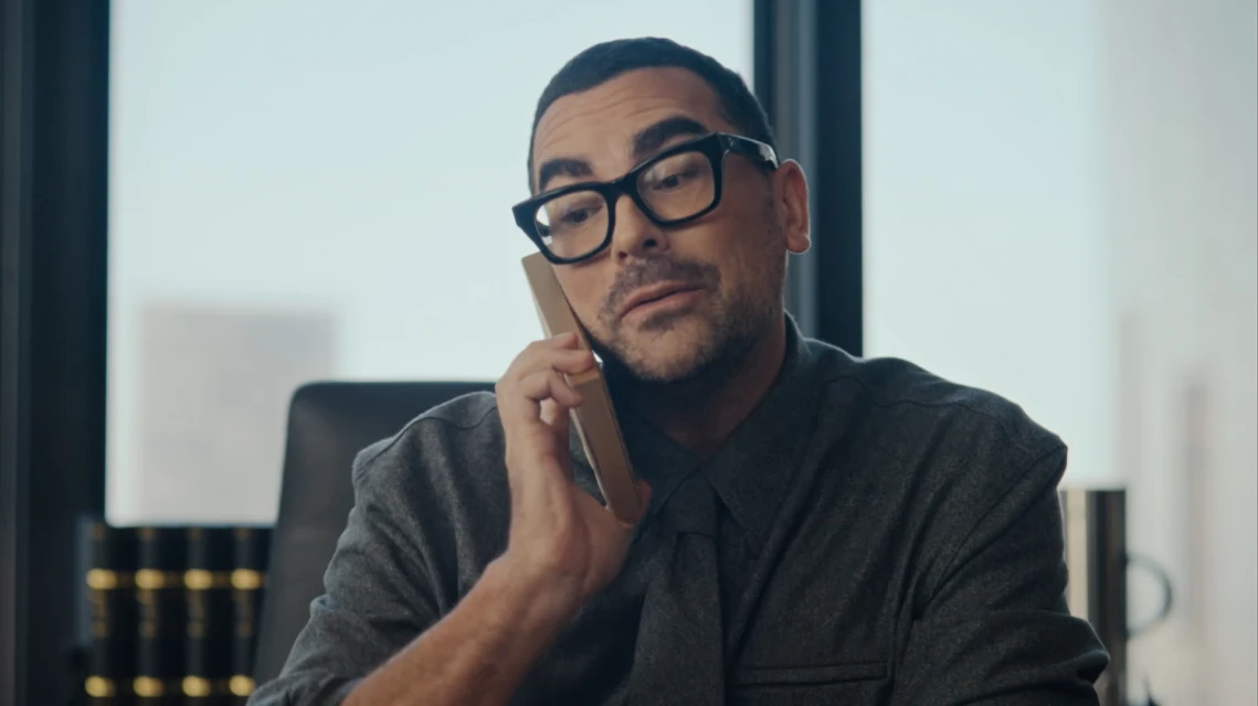 Adult male in black-framed glasses seated at desk, holding up a rectangular phone, in a modern office with a bright window.