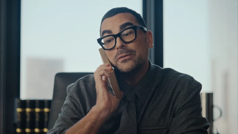 Adult male in black-framed glasses seated at desk, holding up a rectangular phone, in a modern office with a bright window.