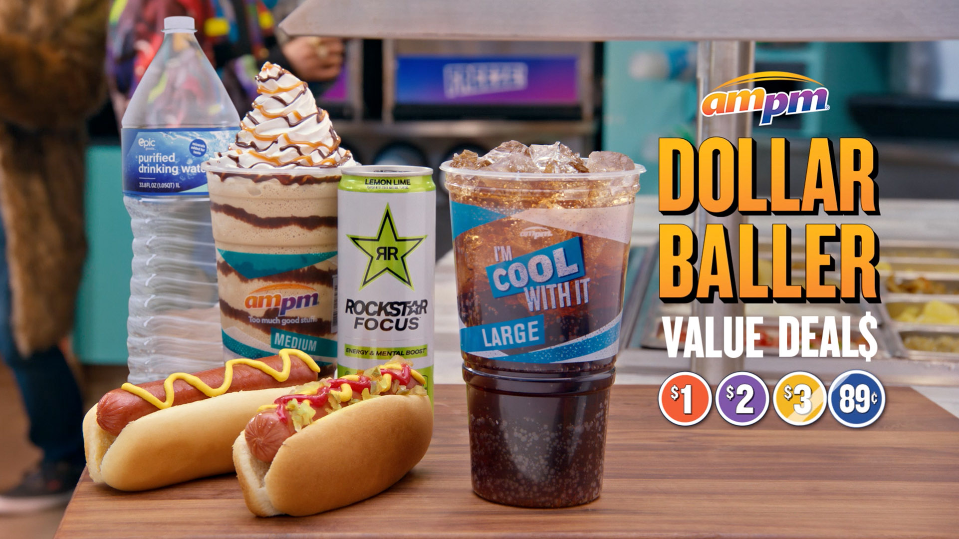 Close-up of two hot dogs, a bottled water, a chocolate milkshake, an energy drink, and a soda with ice on a wooden surface.