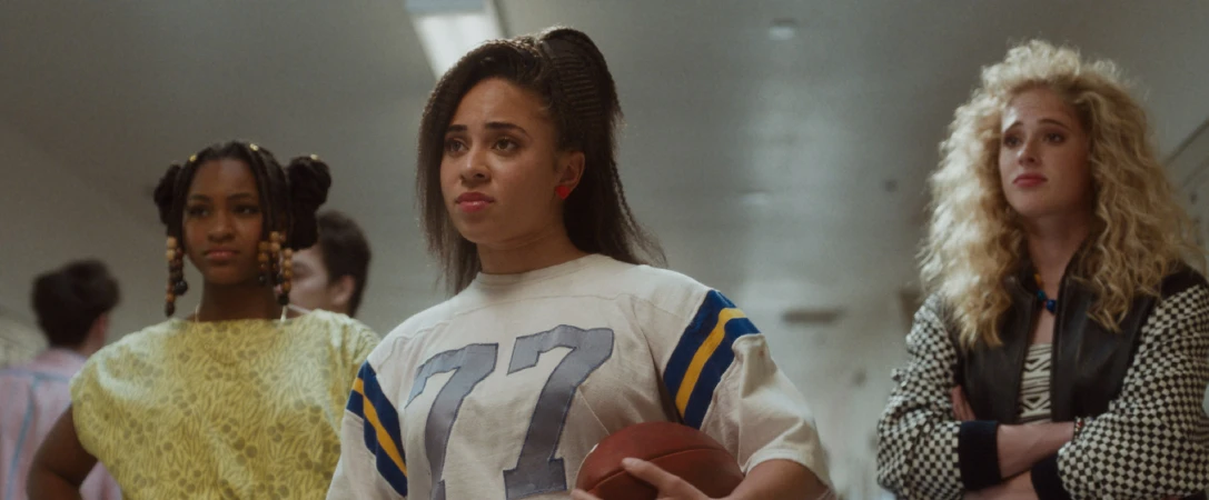 Three young women in vintage clothing stand in a hallway; the central figure holds a football, creating a retro, sporty fashion statement.