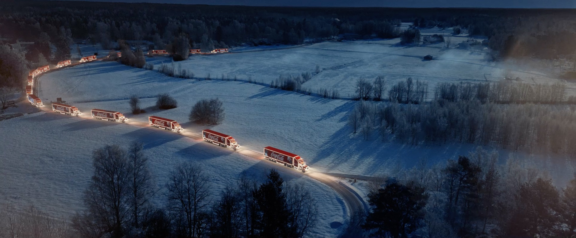 Aerial view of Coca-Cola holiday caravan of trucks on a snowy rural road surrounded by snow covered fields and dark forest.