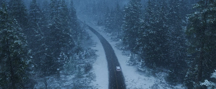 Aerial view of a car driving on a winding asphalt road through a dense snowy forest during a snowfall.
