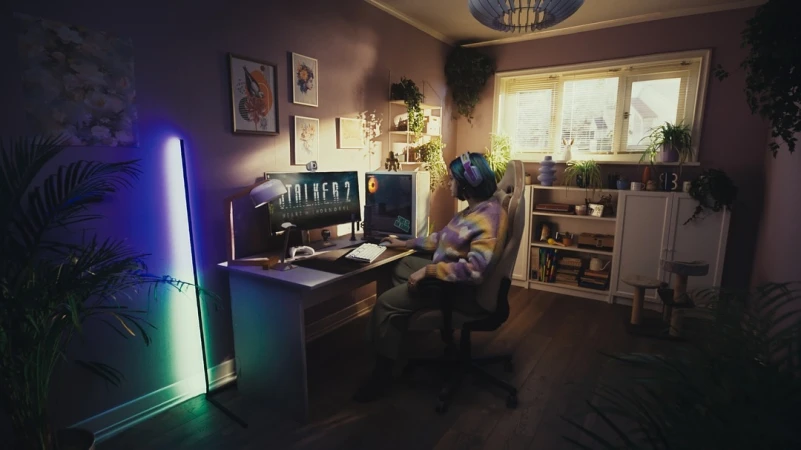A person with blue hair sits in a gaming chair at a desk with dual monitors displaying a video game in a home interior.