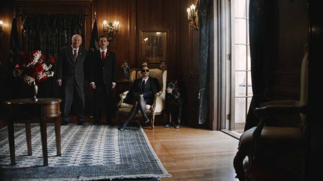 Three people in suits stand in an ornate room with dark wood paneling, a patterned rug, and a dog. One person sits in an armchair.