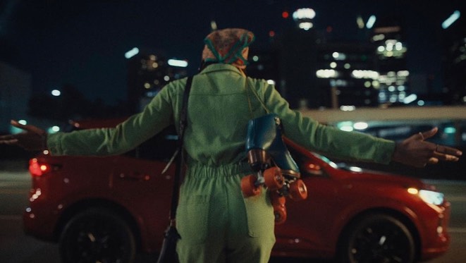An adult with roller skates stands beside a red SUV at night in an urban setting with bokeh lights in the background.