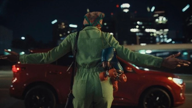 An adult with roller skates stands beside a red SUV at night in an urban setting with bokeh lights in the background.
