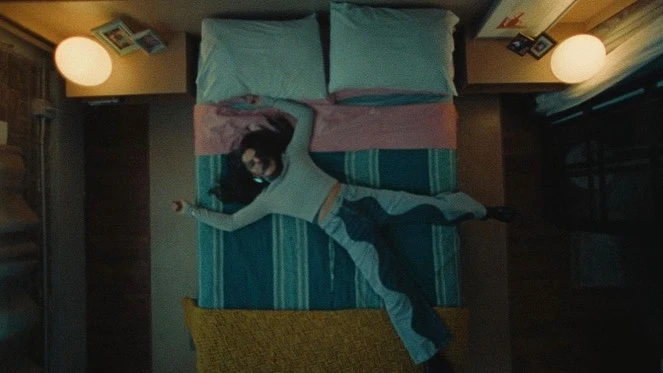 Top-down view of a woman lying flat on a bed with arms outstretched in a dimly lit room directed by Christopher Gruse.