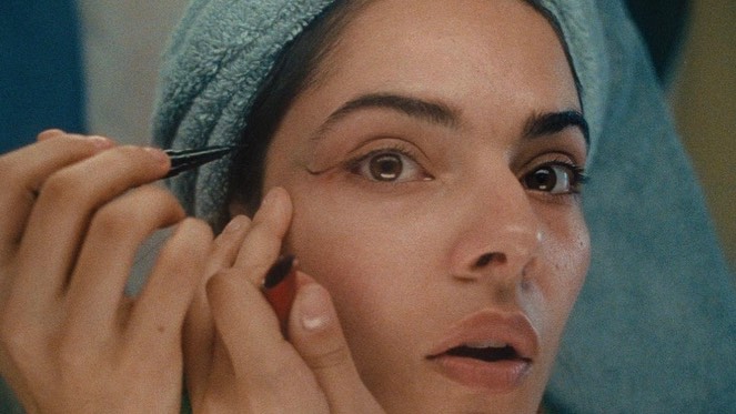 Close-up of a woman applying eyeliner with focused expression, light blue towel around her head, beauty and self-care in soft lighting.