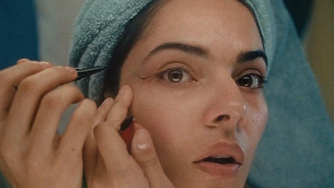 Close-up of a woman applying eyeliner with focused expression, light blue towel around her head, beauty and self-care in soft lighting.