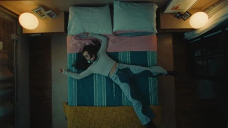 Top-down view of a woman lying flat on a bed with arms outstretched in a dimly lit room directed by Christopher Gruse.