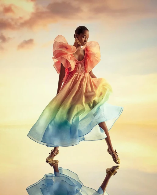 Fashion shoot featuring a stylish Black woman in a flowing, colorful dress on a reflective surface with a dramatic sky backdrop.