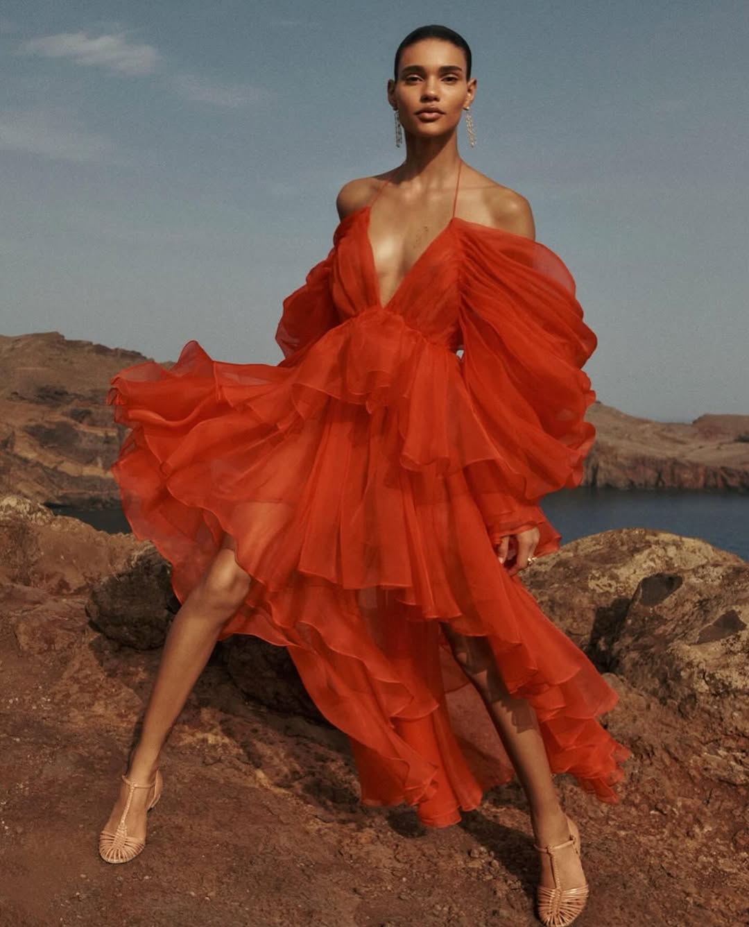 A woman in a red, tiered, off-the-shoulder gown poses on a rocky hillside with ocean and sky background, showcasing fashion and beauty.