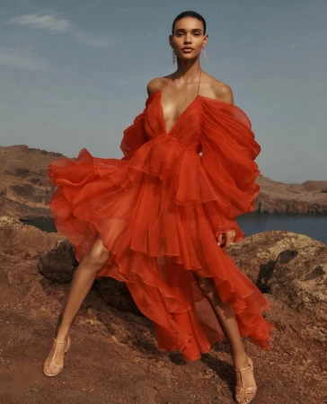 A woman in a red, tiered, off-the-shoulder gown poses on a rocky hillside with ocean and sky background, showcasing fashion and beauty.
