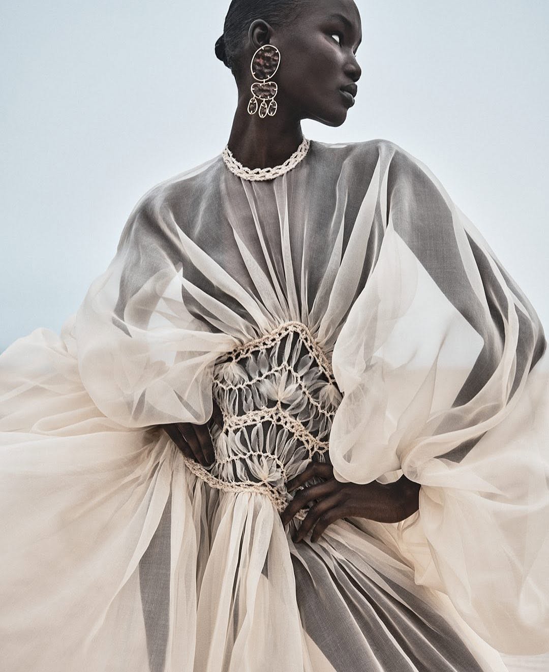 Fashion editorial shot of a Black female model in an elaborate ivory dress with statement earrings and necklace against a pale sky background.