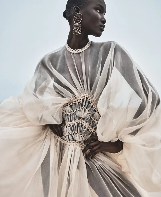 Fashion editorial shot of a Black female model in an elaborate ivory dress with statement earrings and necklace against a pale sky background.