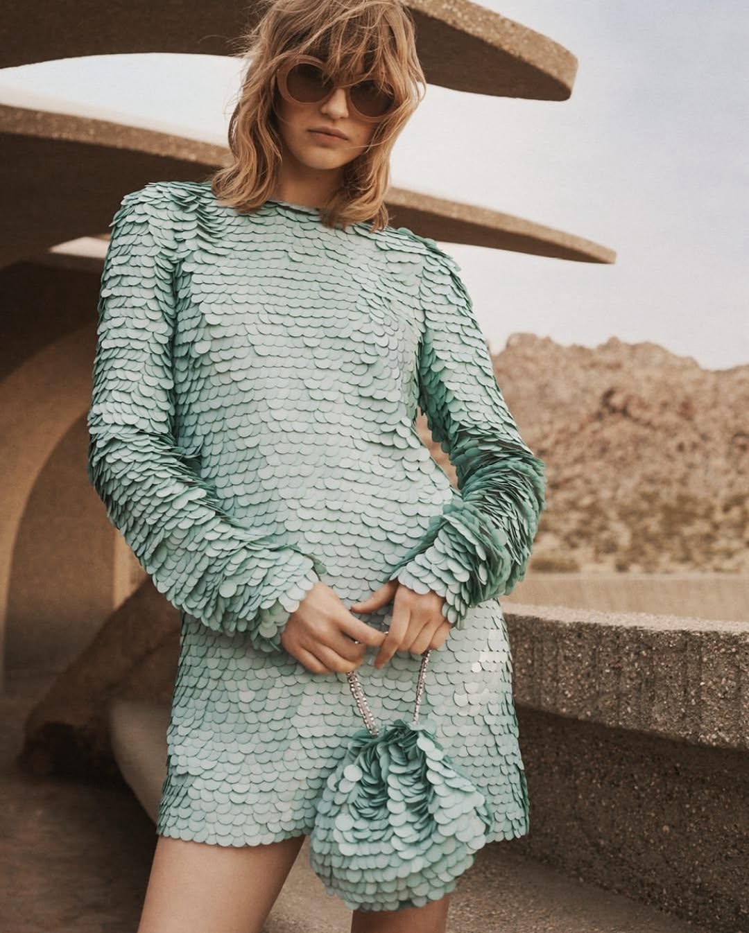 A woman wearing a teal, sequined dress and round sunglasses stands on a terrace in front of a desert landscape on a sunny day.