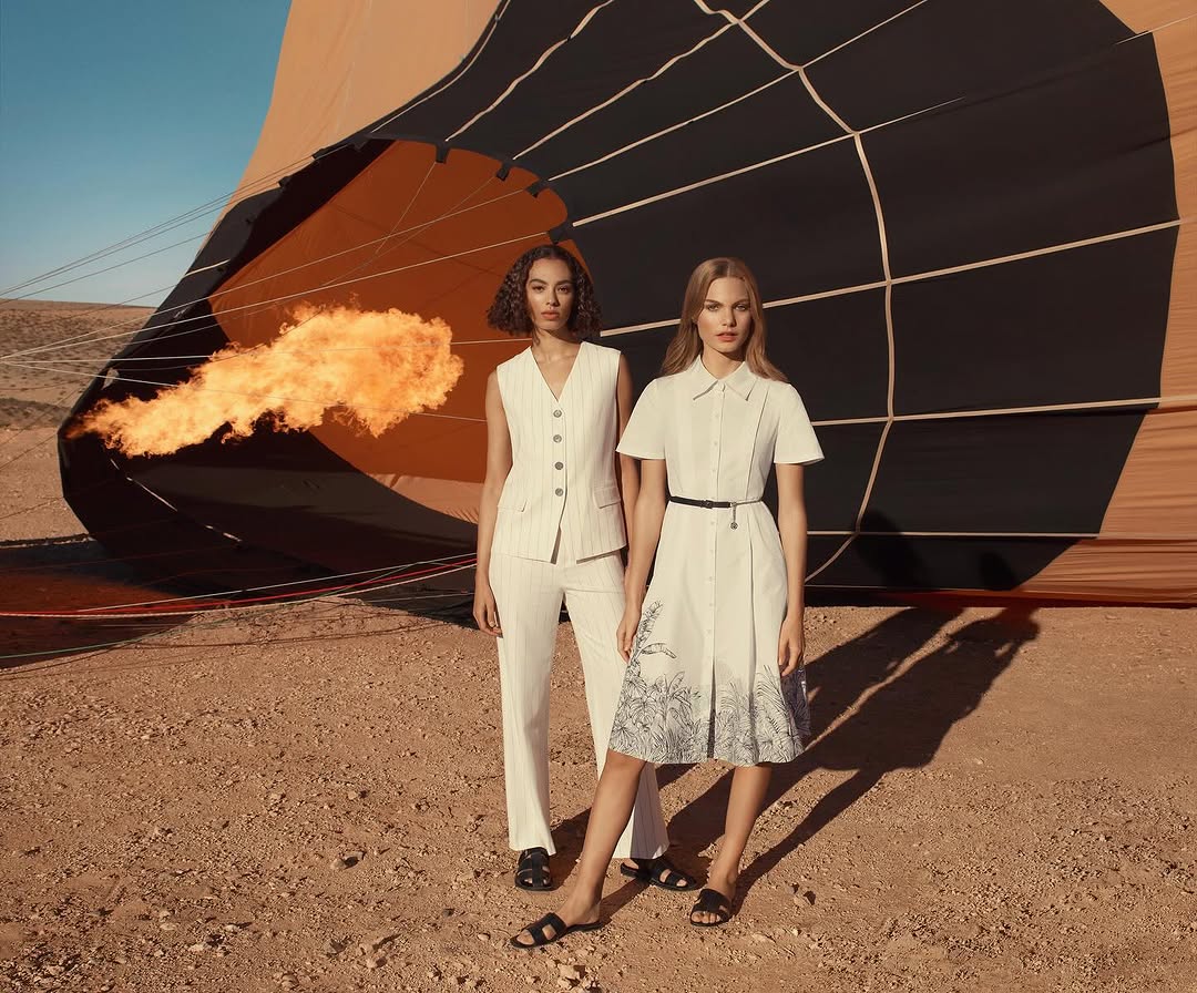 Two women in fashionable outfits pose in a desert landscape with a hot air balloon and a burst of flame in the background.