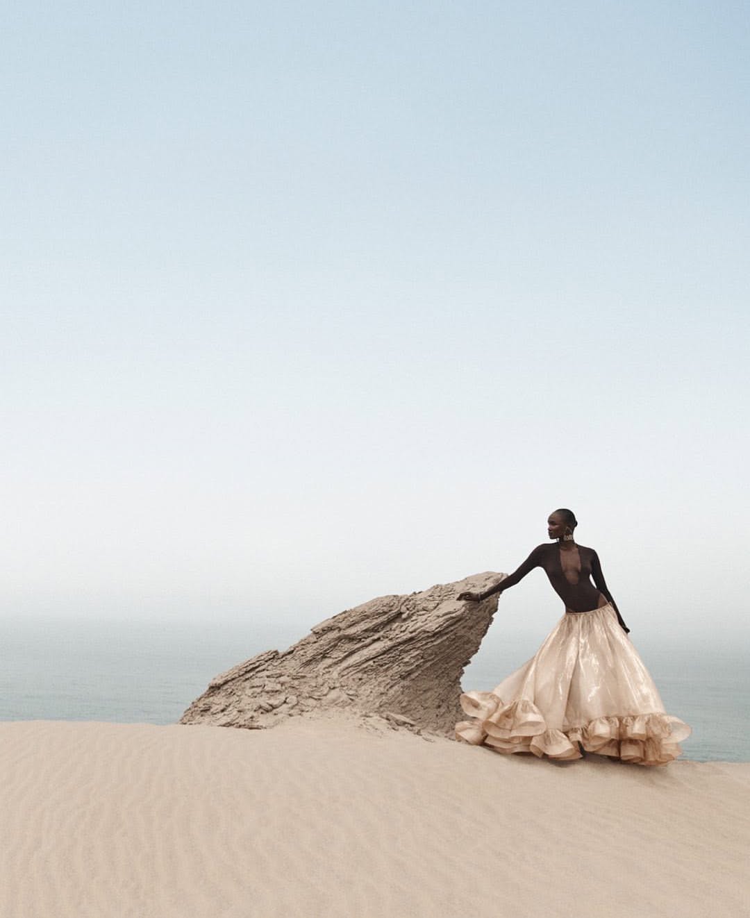 A dark-skinned model in a flowing beige dress poses on a sand dune with a rock formation, ocean, and sky in the background.