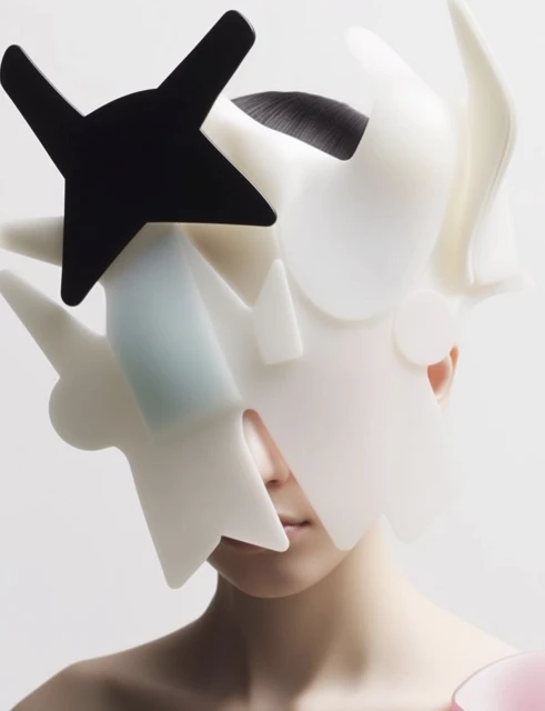 Artistic close-up shot of a model with a sculptural headpiece featuring white and black abstract geometric shapes against a white backdrop.