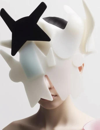 Artistic close-up shot of a model with a sculptural headpiece featuring white and black abstract geometric shapes against a white backdrop.