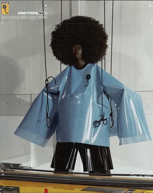 Fashion-forward portrait of a Black model with an afro, wearing a glossy blue oversized top and black shorts in a modern setting.