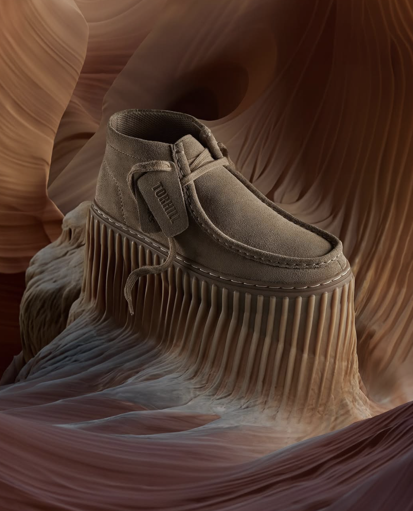 Beige suede chukka boot with fringe detail on a textured brown and beige sculptural surface, studio shot, neutral color palette.