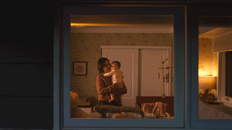 A woman holding a baby inside a dimly lit nursery seen through a window from the exterior at night.