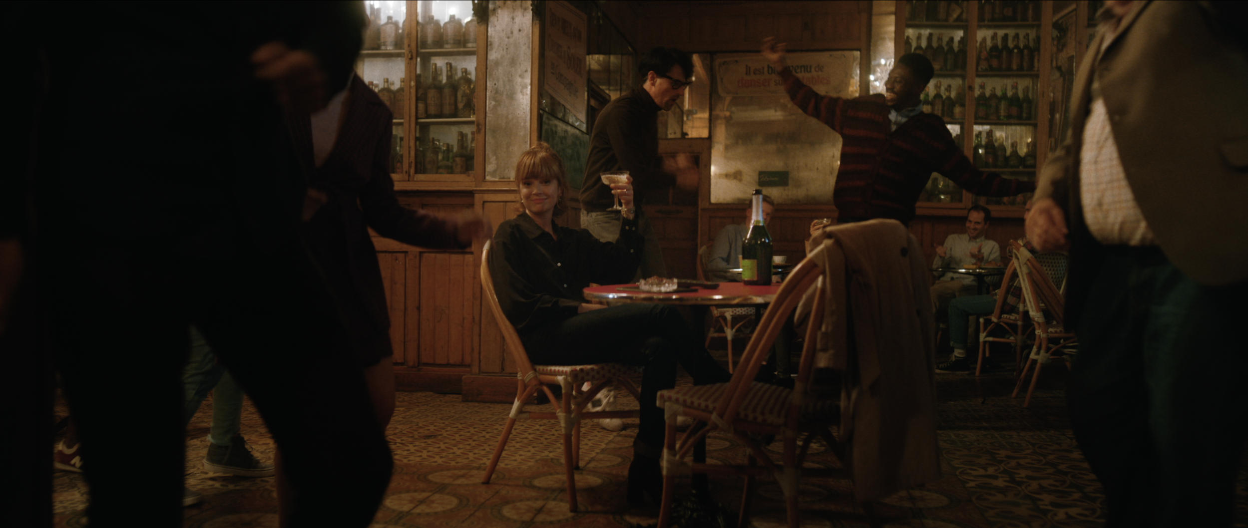 Woman sits with cocktail at dimly lit bar as others dance in the background. Wood paneling and patterned floor create an intimate atmosphere.